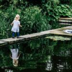 A young girl stands on a bridge overlooking a serene pond, surrounded by lush greenery and gentle reflections in the water.