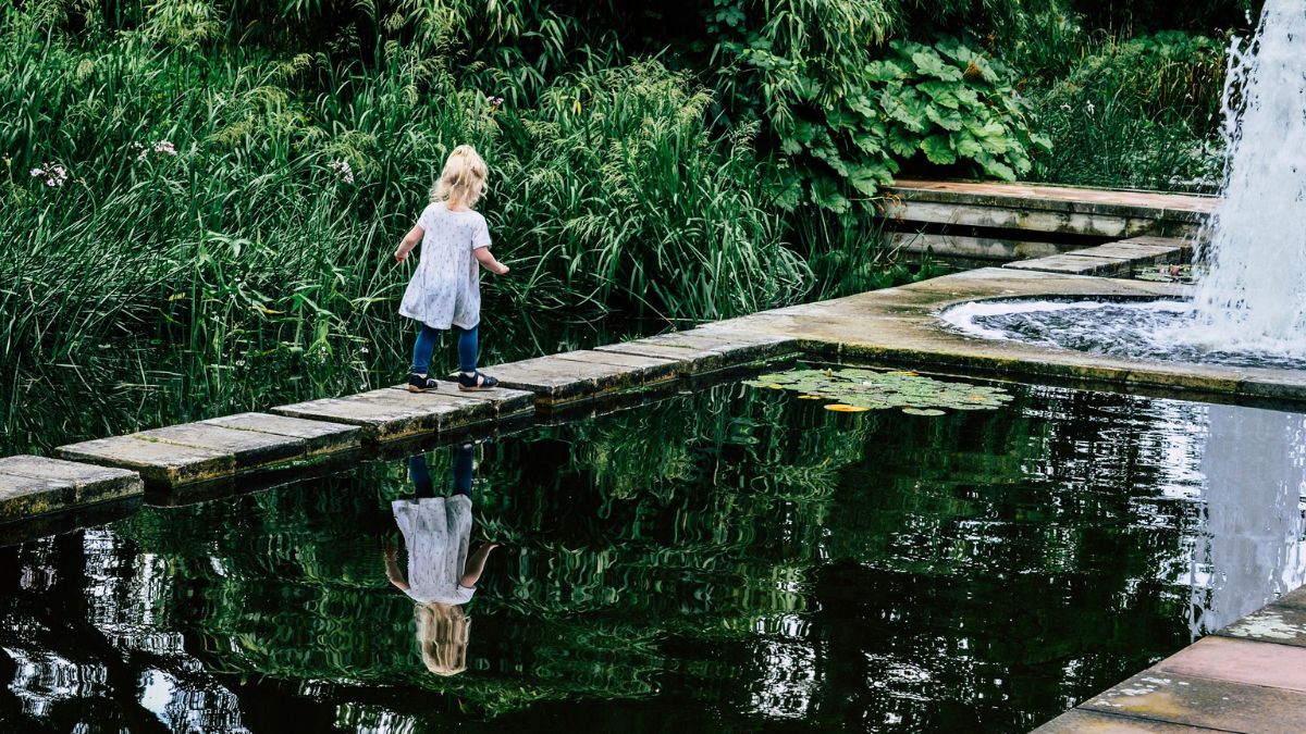 A young girl stands on a bridge overlooking a serene pond, surrounded by lush greenery and gentle reflections in the water.
