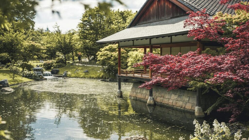 A serene Japanese garden featuring a tranquil pond and a traditional house surrounded by lush greenery.