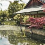 A serene Japanese garden featuring a tranquil pond and a traditional house surrounded by lush greenery.