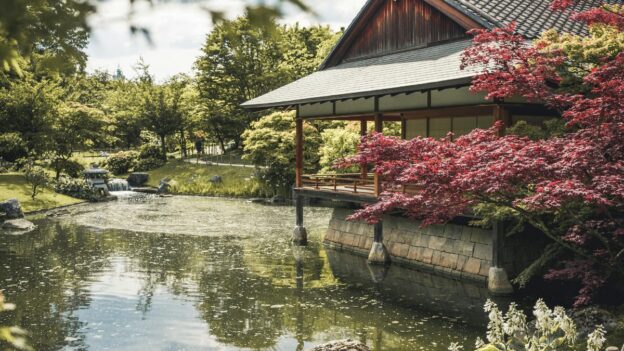 A serene Japanese garden featuring a tranquil pond and a traditional house surrounded by lush greenery.